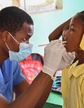 A dentist in blue scrubs, mask & gloves peers into the mouth of a girl in a yellow school uniform and hair ribbons.