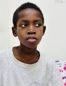 A young Haitian boy with short dark hair sits on a hospital bed. He looks off to the right side.