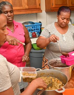 Three Haitian women stand around a kitchen table, spooning white rice, black bean sauce, and mixed vegetables into colorful single-serving containers.