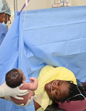 In an operating room, a nurse in full PPE holds out an infant to her joyful mother who is still on the operating table, under drapes.