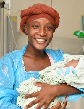 •	A young Haitian woman wearing a orange-brown head wrap and blue hospital gown smiles happily. She holds a small infant in her arms.