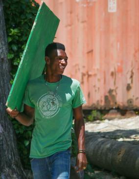 A Haitian man wearing a green T-shirt and blue jeans looks over his shoulder and smiles. He is carrying a piece of green-painted plywood.