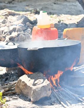 Two large pots sit on cinderblocks and rocks above an open fire in a sunny yard. One pot is covered, and the other has steam rising from it.