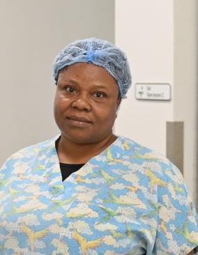 A woman Haitian nurse anethesist stands in front of a doorway wearing light blue scrubs and a blue hairnet. She smiles at the camear.