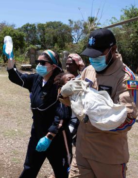 A uniformed male EMT carries a baby swaddled in white across a dry dirt field. Beside him walk two women, one holding up an IV bag.
