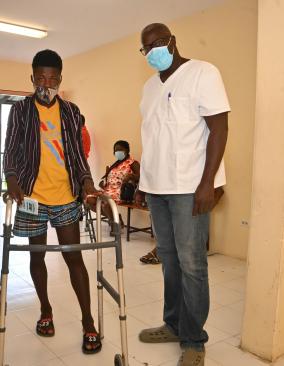A young man stands, holding a walker, next to an older, taller man wearing glasses and a white scrub shirt. Both wear face masks.