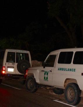 Three white ambulances drive in a row with their headlines on through pitch-black night.