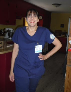 A light skinned woman stands in a wood-framed doorway dressed in dark blue medical scrubs. She has one hand on her hip and smiles at the camera.