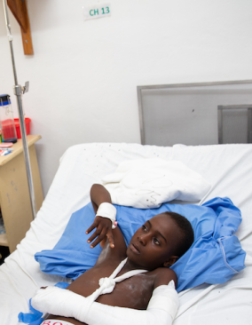A Haitian boy likes in a hospital bed; his right arm is bandaged up to his elbow. A Haitian man leans over his bed to talk to him.