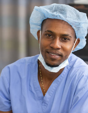 Headshot of a Haitian doctor leaning forward, smiling in blue scrubs, surgical cap & pulled-down surgical mask.