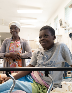 A young Haitian boy props himself up in a hospital bed. He smiles at the camera. His mother stands behind him with her hands together, also smiling.