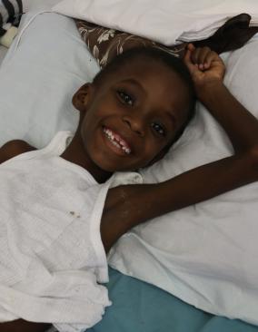 A young Haitian boy lays on a hospital bed in a white tank top. He smiles and holds his arm above his head.