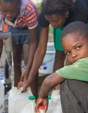 Haitian children fill water jugs at a pump. A boy turns his head toward the camera; 2 older girls focus on the water.