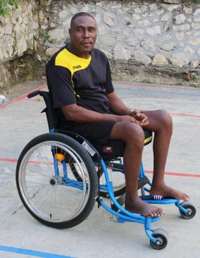 A Haitian man sits in a wheelchair on a concrete basketball court. He turns his head to the side to look at the camera.