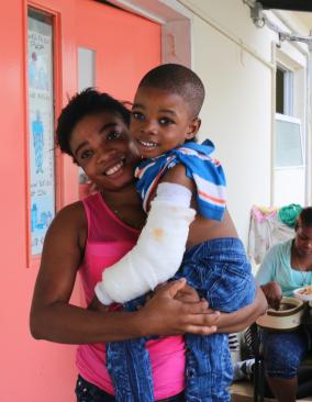 A Haitian woman holds her young son in her arms. Her son's arm is in a cast up to his elbow. They both smile.