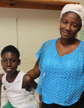 A young Haitian boy sits on a hospital bed and smiles. His mother stands next to him, with her arm around his shoulder.
