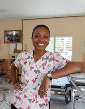 A Haitian physical therapist wears short-sleeve scrubs and stands smiling in front of physical therapy equipment.