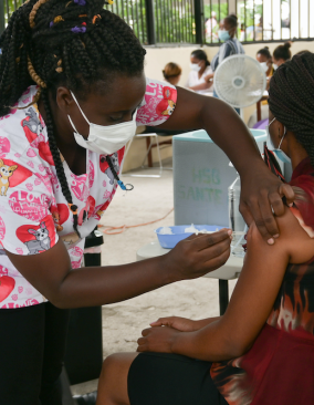 A nurse in scrubs with pink & red hearts administers a COVID-19 vaccine. Both women are Haitian and have long braids.