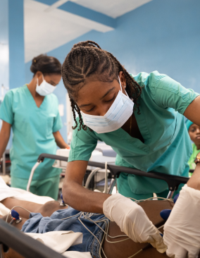 A young female Haitian clinician wearing a medical mask bends over a patient in a hospital bed, adjusting some sensors on the patient's side.