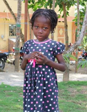 A young Haitian girl wearing a polka-dot dress stands in front of a gravel path lined with trees. She clasps her hands to her chest.