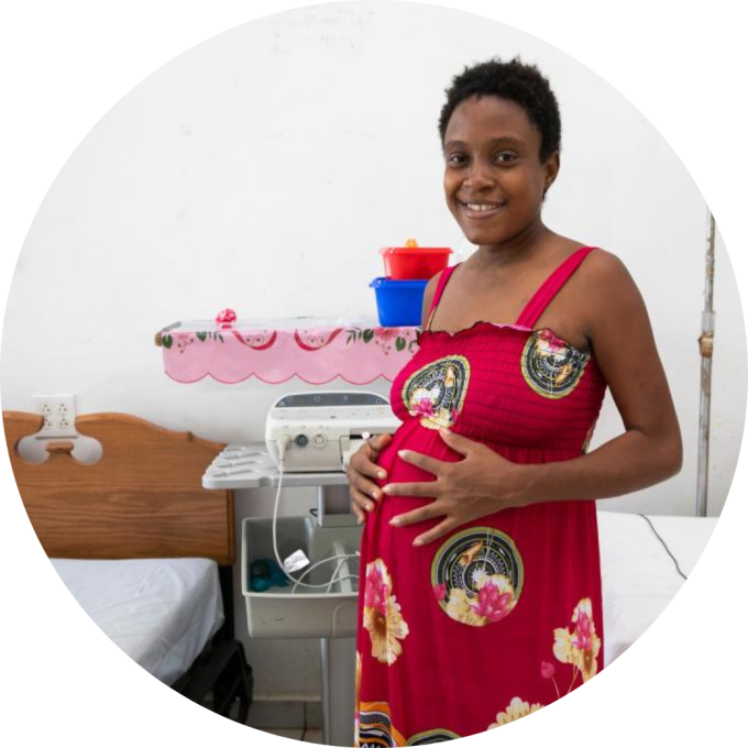A pregnant Haitian woman wearing a sleeveless red floral dress touches her stomach and smiles.