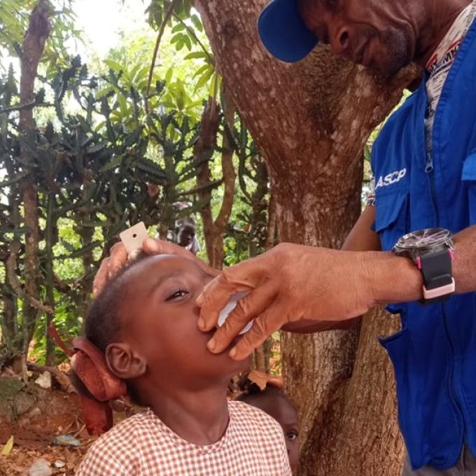 In the Haitian countryside, a man wearing a bright blue cap and a vest with official logos holds a small cup up to a young boy’s mouth.