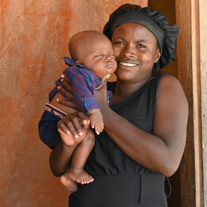 A Haitian woman stands in a wooden doorway. She holds her infant son in her hands, held up near her face. He wears blue shorts and a plaid button down-shirt. He is sleeping. The woman wears a black hair bonnet and a sleeveless black dress. She has a bright smile.