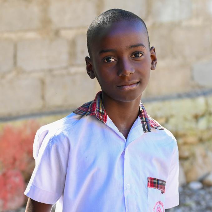 A young Haitian boy stands in front of a cinder block wall and gives a small smile to the camera. He has very short black hair and wears a white short-sleeve button-down shirt with a red plaid collar and pocket.