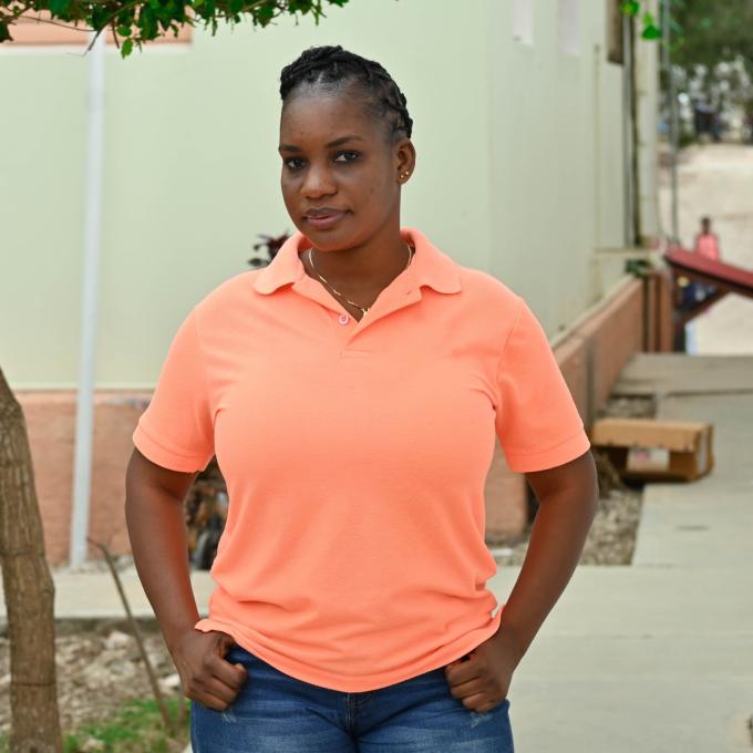 A Haitian woman community health worker stands in front of a building and gives a small smile at the camera. She wears a coral-colored polo shirt. Her hands are on her hips, and her hair is intricately braided.
