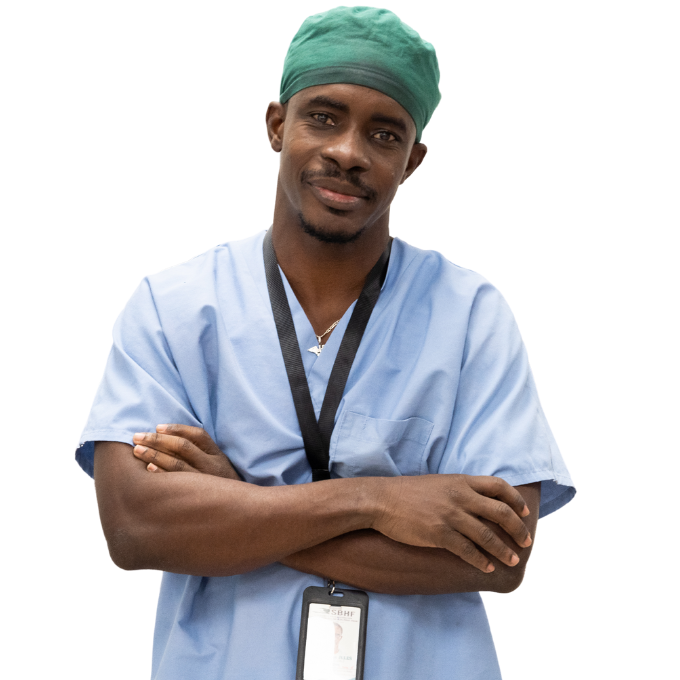A Haitian surgeon stands with his arms crossed looking directly at the viewer. He wears light blue scrubs, a black nametag lanyard, and a tight turquoise-colored surgical cap.