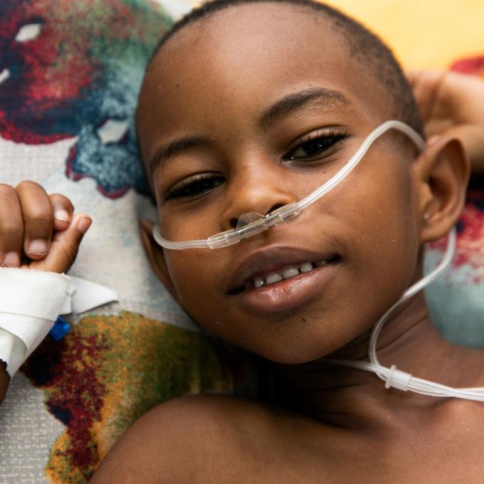 Close-up portrait of a young Haitian boy smiling at the camera. As he relaxes on a patterned blanket, his hands by his head, tubes bring oxygen to his nostrils.