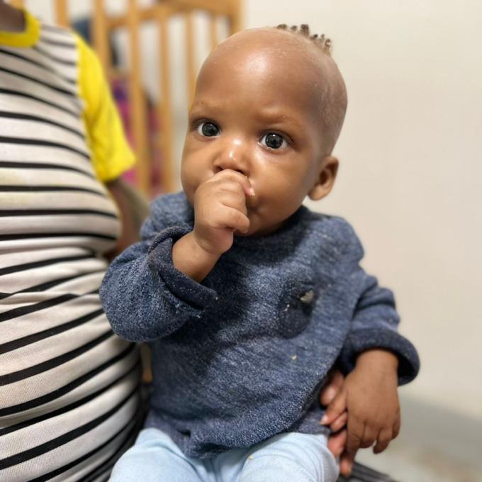 A young girl Haitian baby sits in her caregiver's lap. The baby has big, bright brown eyes and only a light dusting of light brown hair. She wears a dark blue sweater and sucks her thumb. Her caregiver's black and white striped shirt is seen to the left.