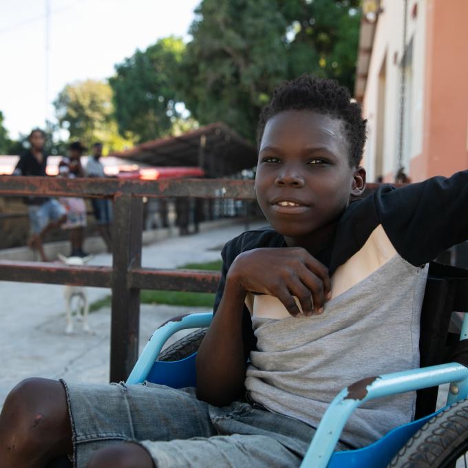 A Haitian teen sits in a bright blue wheelchair. His upper body is turned to face the camera. He is smiling.
