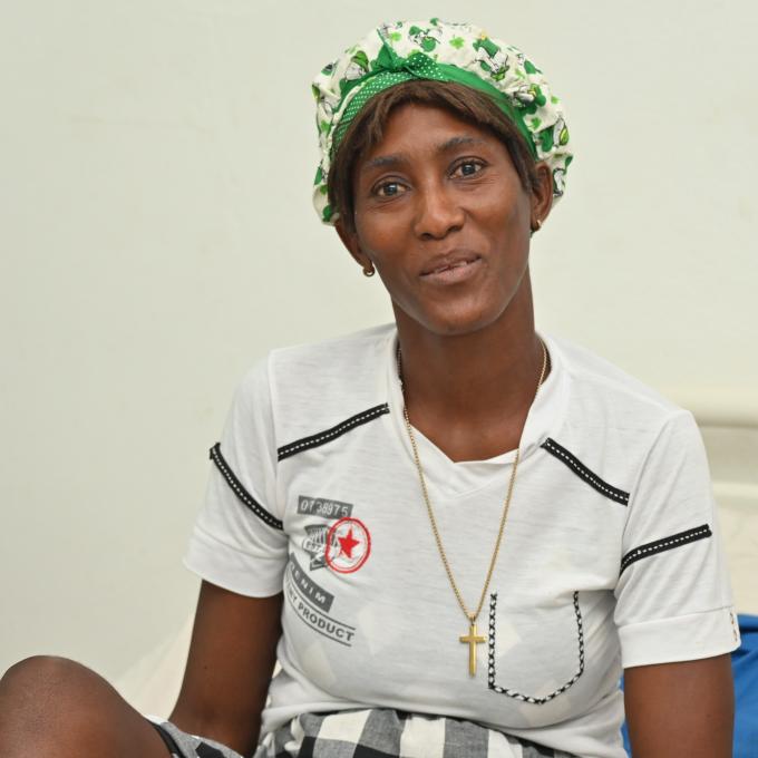 Portrait of a Haitian woman smiling gratefully at the camera. She wears a green head wrap, a white knit shirt, and a large gold cross.