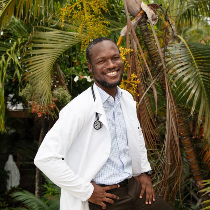 A Haitian male doctor stands in front of palm trees, smiling brightly at the camera. He wears a white doctor's coat and has a stethoscope around his neck.