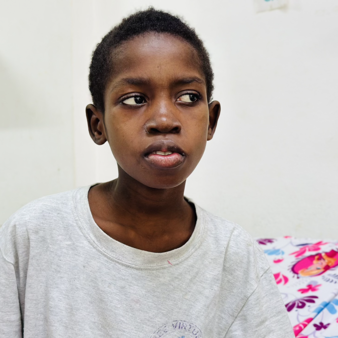 A young Haitian boy with short dark hair sits on a hospital bed. He looks off to the right side.