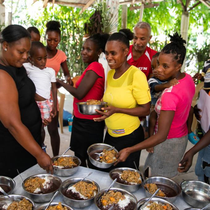 A dozen Haitian adults and small children in bright colors gather around a table, smiling. The mothers reach for metal bowls full of cooked food.