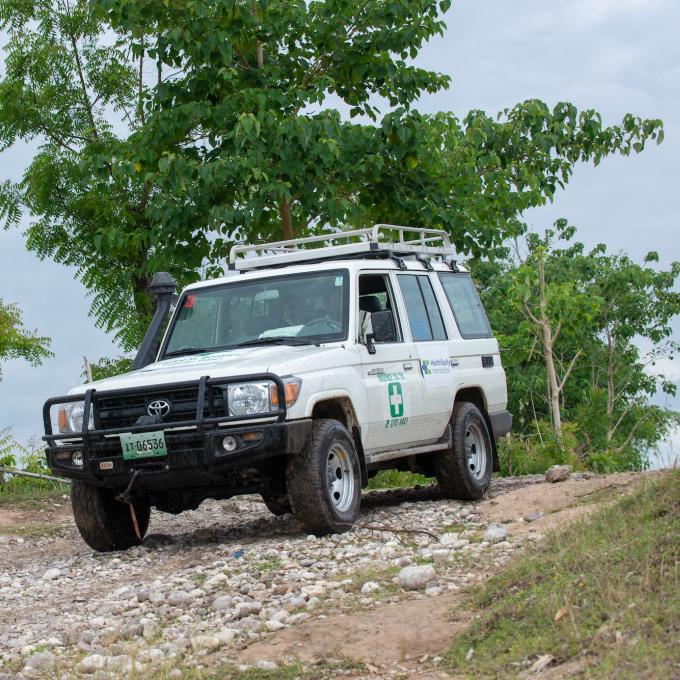 A white SBH ambulance drives along a bumpy country road. The road is unpaved and slopes downward slightly, from right to left. The sky is blue and cloudy. The car is driving in front of some low green trees and brush.