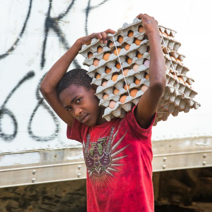 A young Haitian man carries stacks of egg cartons over his left shoulder. His arms are above his head securing the bundle. He wears a red t-shirt.