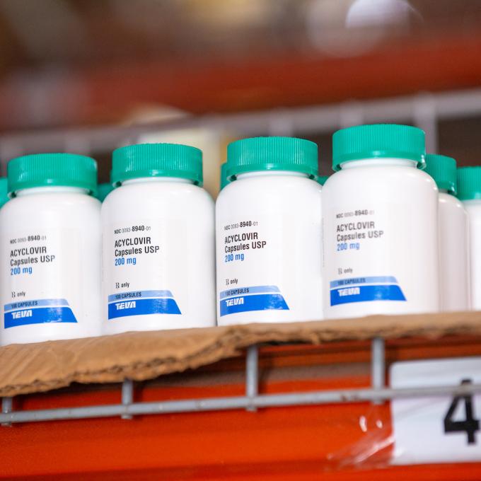 Several bottles of the same kind of medicaton stand on a wire warehouse shelf. The bottles are small and made of white plastic. The white label has black text on it with a dark blue bar across the bottom. The bottle cap is a bright teal.