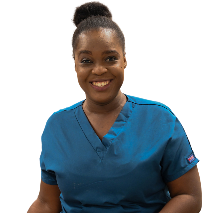 A Haitian nurse is shown from the waist up. She has a big smile. She wears dark blue short-sleeved scrubs and her hair up in a bun 
