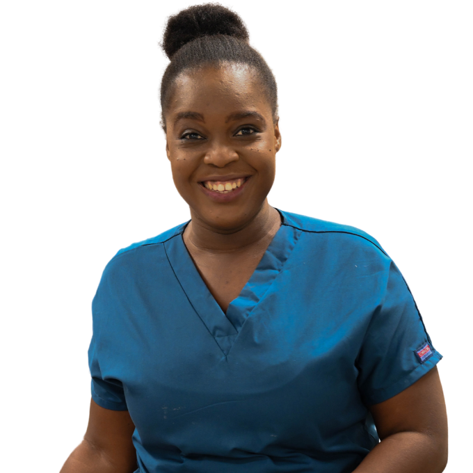 A Haitian nurse is shown from the waist up. She has a big smile. She wears dark blue short-sleeved scrubs and her hair up in a bun