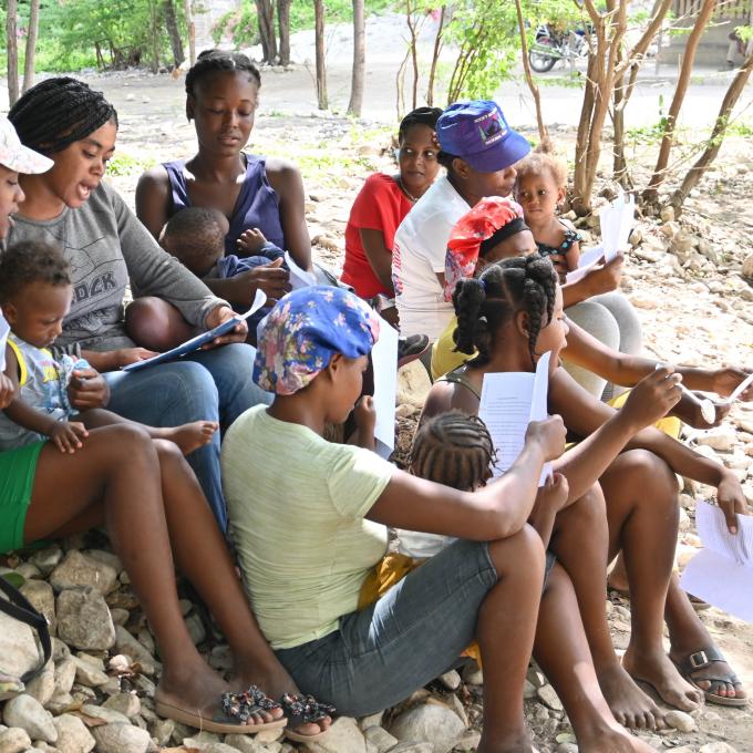 A group of mothers and their children sit on the ground and sing a song about nutrition.