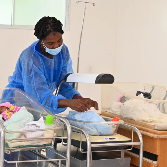 In a hospital, a Haitian woman in a blue gown and surgical mask arranges a blanket on a newborn situated in a bassinet. Two additional newborns rest, one on each side of her.
